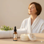 Huile d’argousier et beurre hydratant Argola au premier plan, avec une femme en peignoir floue en arrière-plan, atmosphère douce et apaisante pour peaux sensibles.